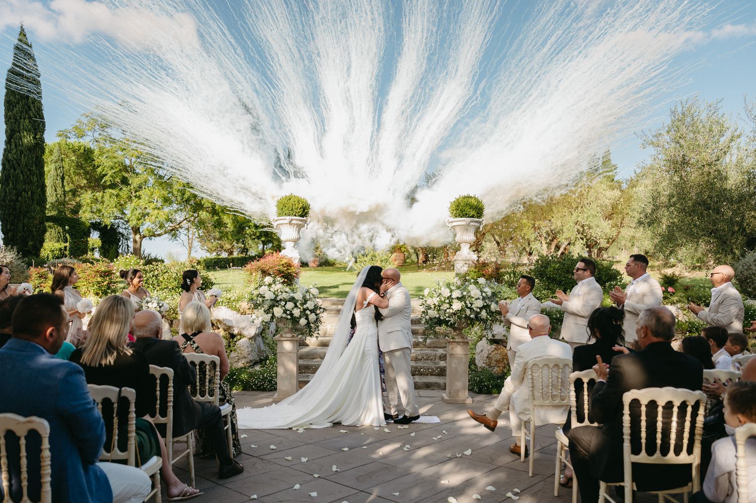 Wedding guests seated outdoors under white draped canopies during a garden ceremony.