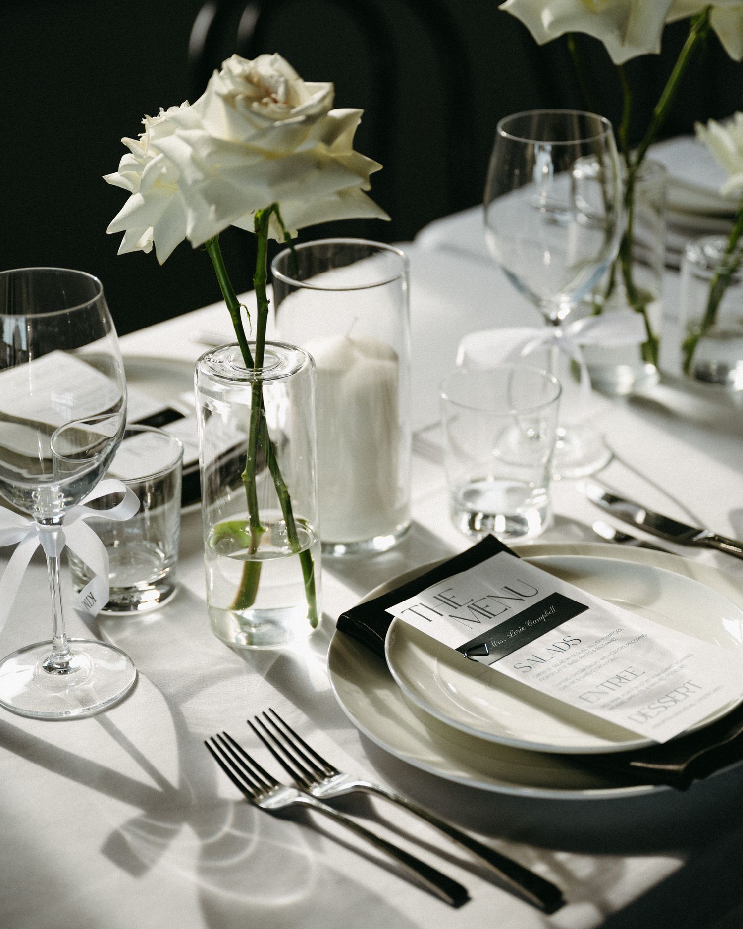Elegant table setting with white roses, wine glasses, and menu cards on crisp white linens in dramatic lighting.