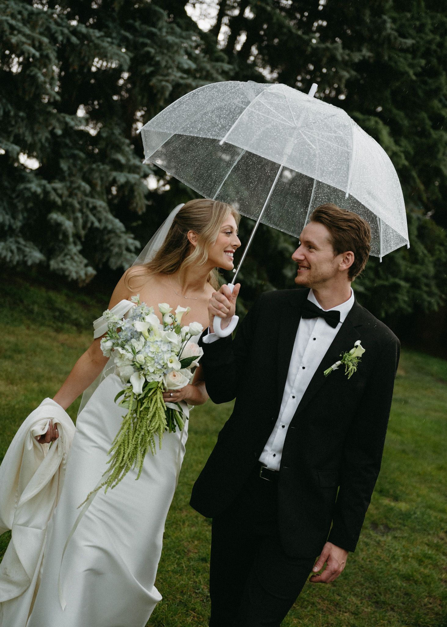 A couple shares a clear umbrella while dressed in formal wedding attire on a rainy day outdoors.