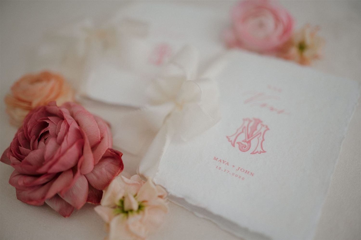 White monogrammed wedding invitation with pink roses and floral decorations on soft neutral background.
