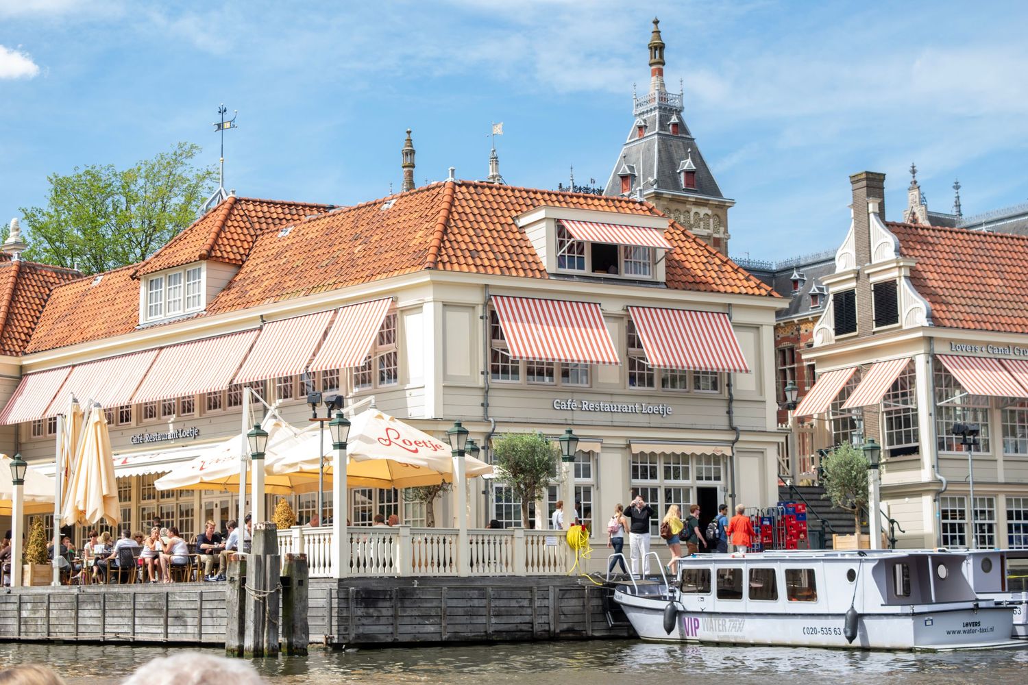 Waterfront restaurants and shops with outdoor seating along a canal in Amsterdam with moored boats.