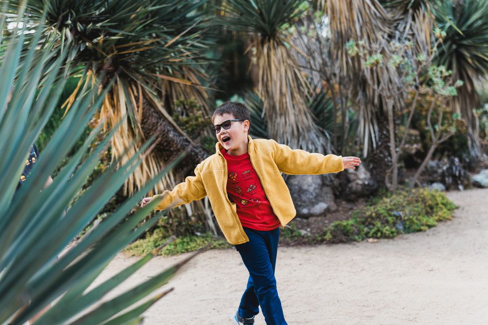 Child in yellow jacket and blue pants poses expressively among desert plants.