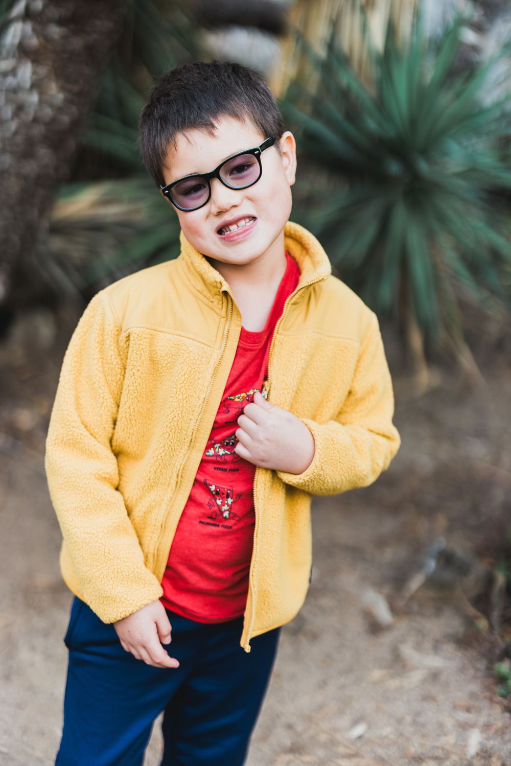 Child wearing glasses, yellow jacket and red shirt smiles while standing in a garden with palm trees.