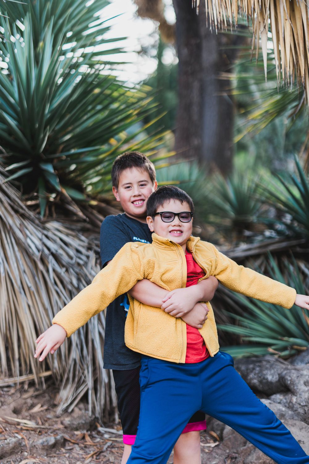 Two children play and laugh together in front of palm trees and desert plants.