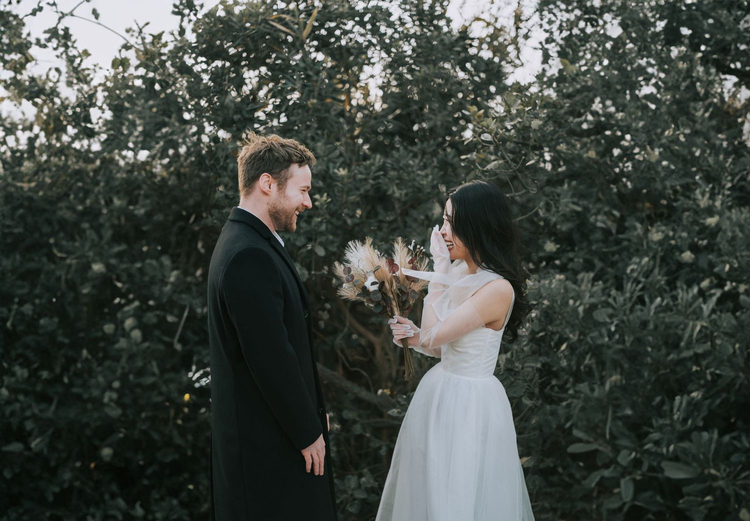 A couple standing in front of greenery, smiling at each other on their wedding day