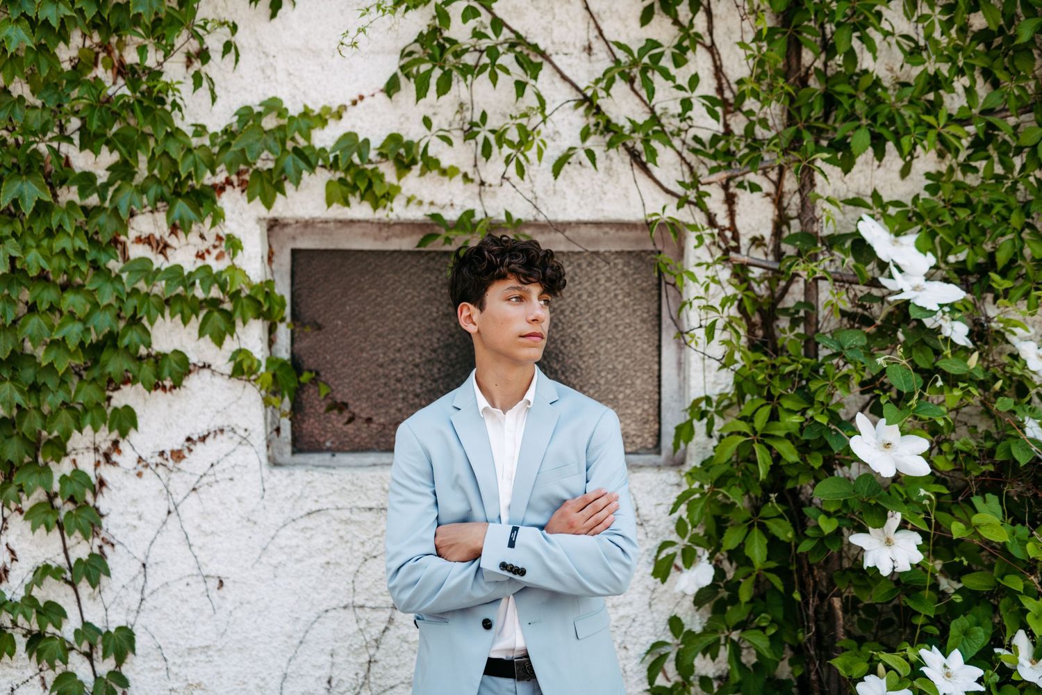 Stylish figure in blue suit posed against white wall with green climbing vines.