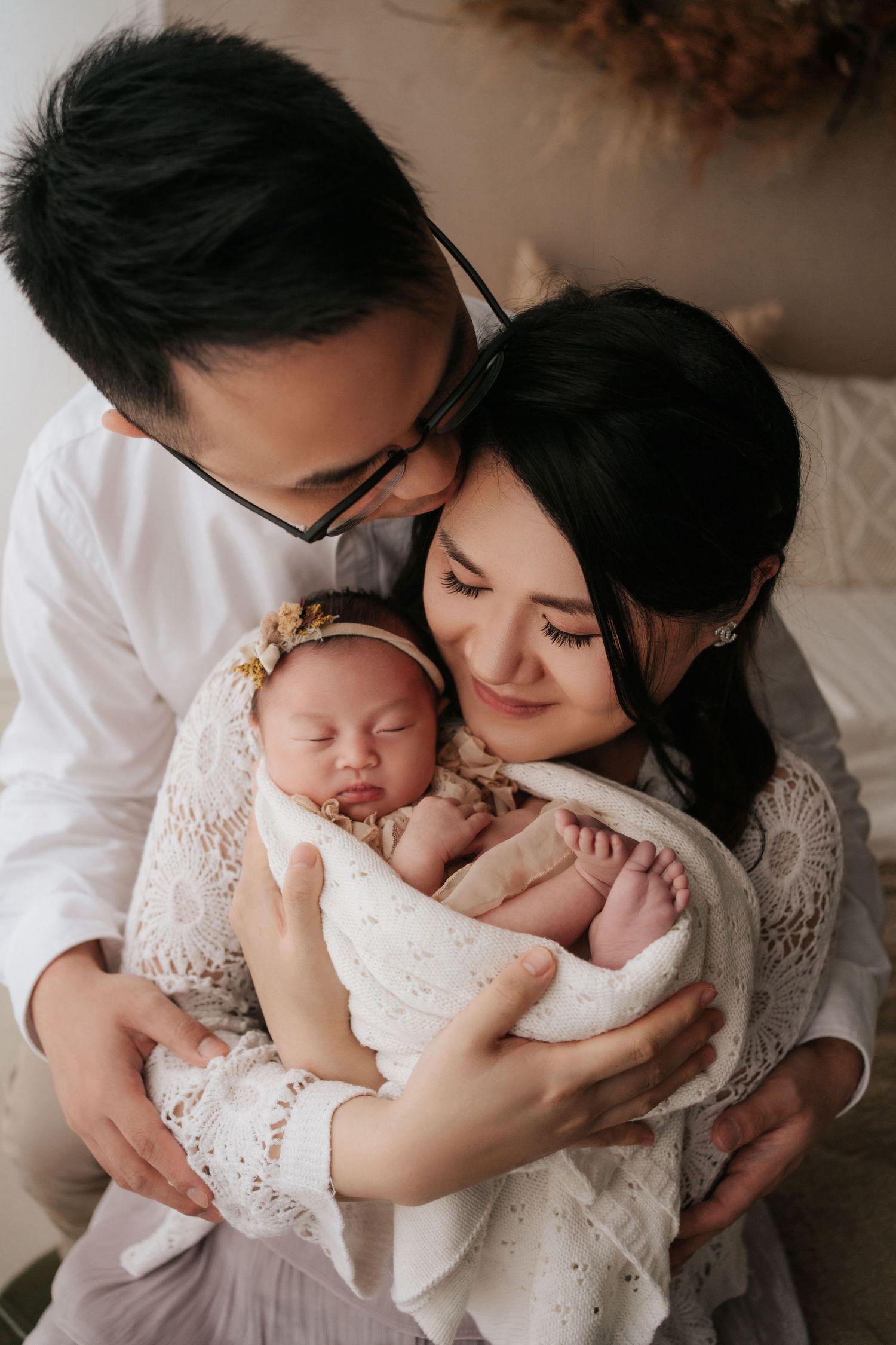 A tender family moment as parents cradle their sleeping newborn baby wrapped in white lace.