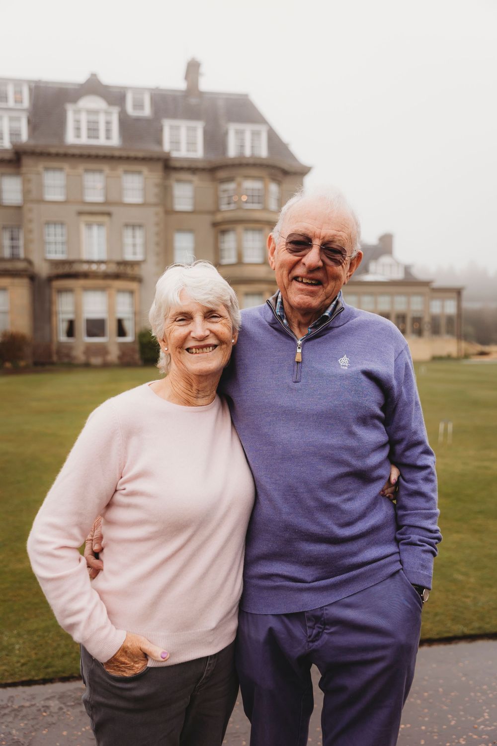 Elderly couple wearing pink and purple sweaters embrace and laugh outside a stately brick building.