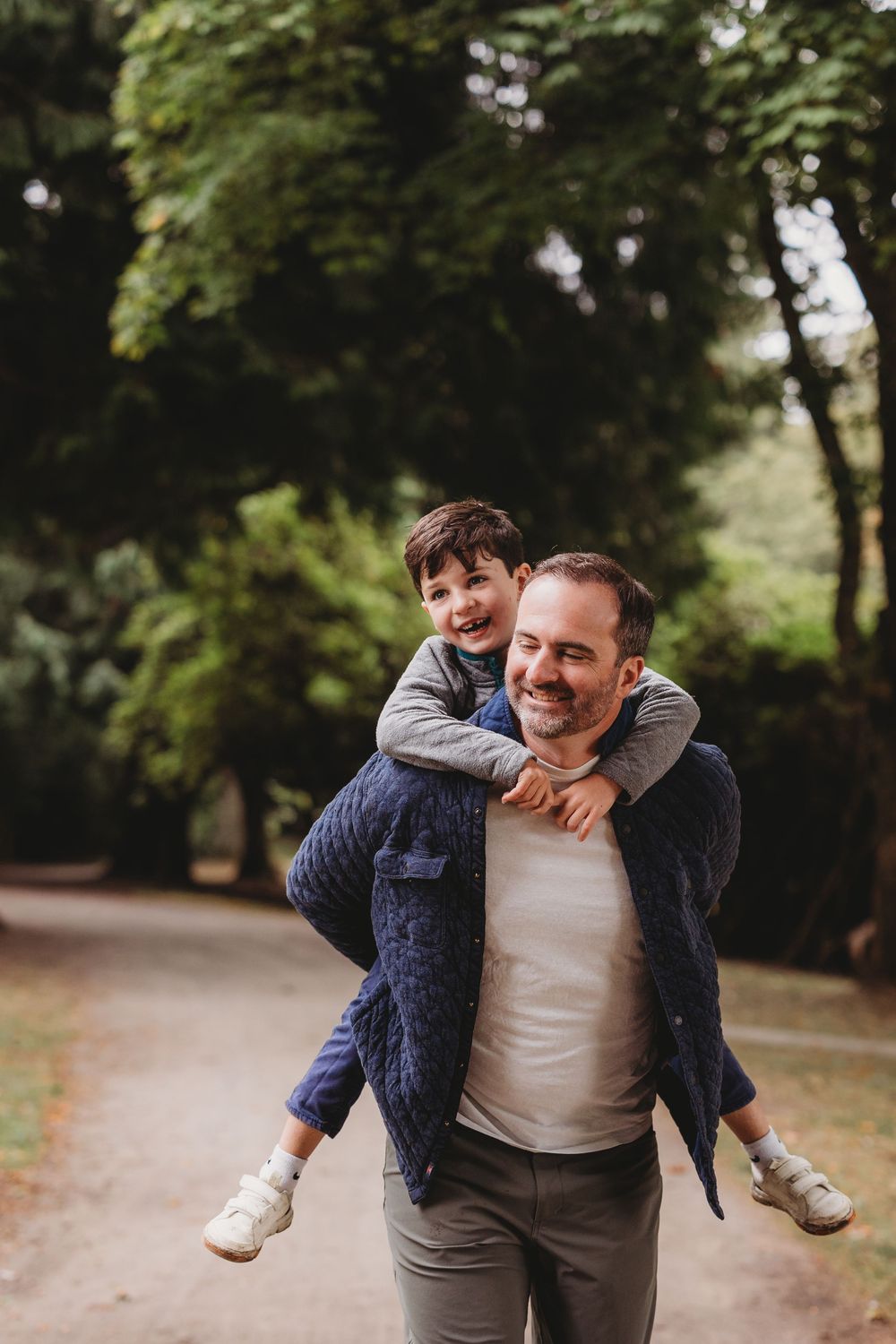 Candid playful father-child moment captured in lush green park path showing joyful laughter and bonding.