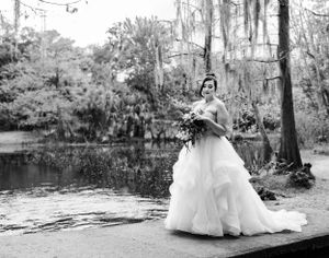 Black and white photo of a bridal portrait in a flowing white gown by a scenic pond surrounded by moss-draped trees.