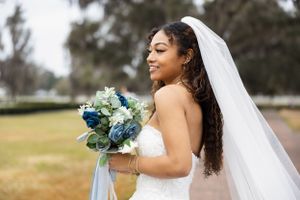 A bride holds a blue and white bouquet while wearing a flowing veil outdoors.