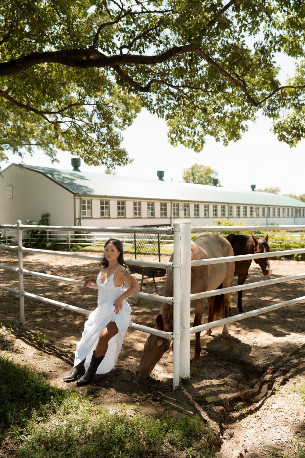 Girl in a white dress standing in front of a fence at a horse stable, with horses visible in the background