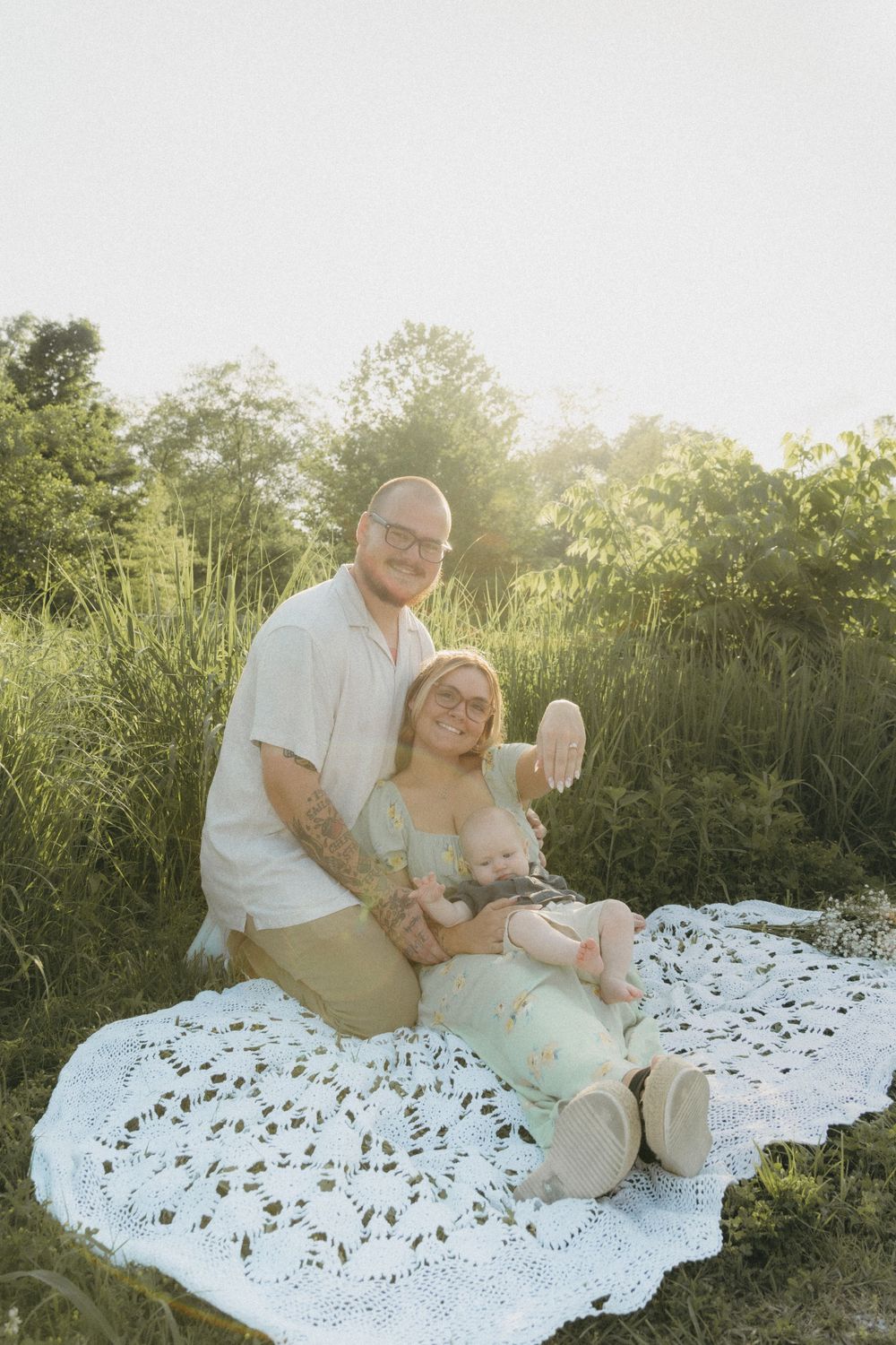 Newly engaged couple sitting on a blanket with their 1-year-old baby, all smiling and enjoying the moment together.