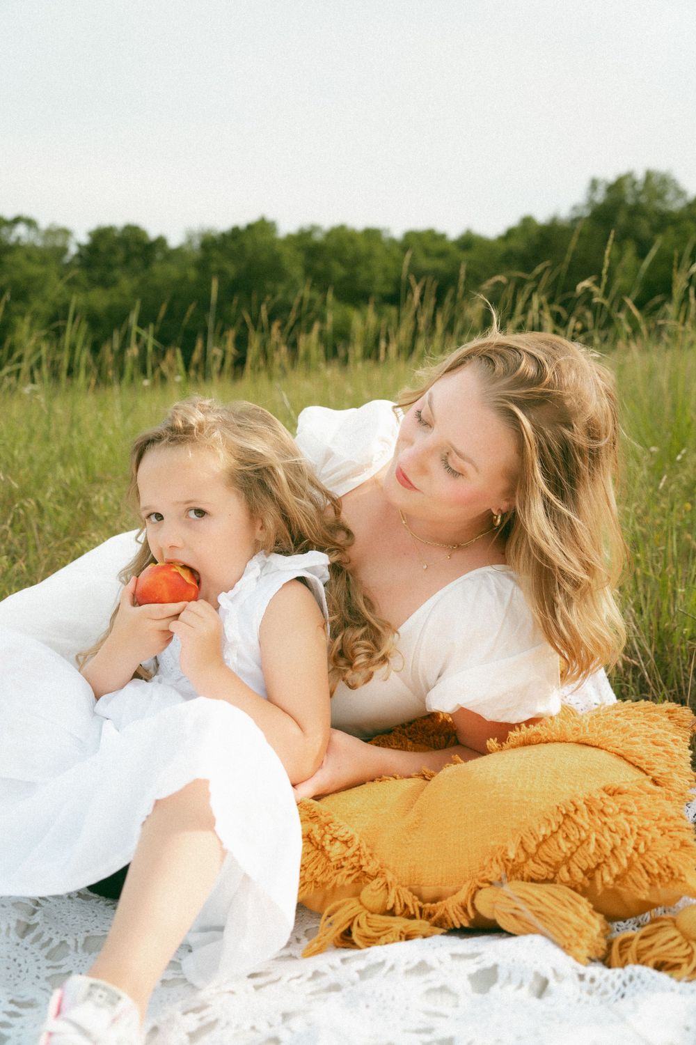 Mom and daughter sitting on a blanket outdoors, with the daughter enjoying a juicy peach while the mom smiles at her.