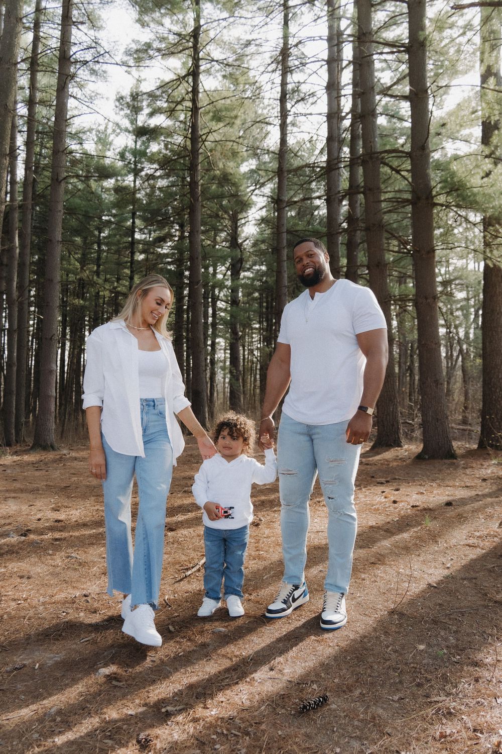 Couple in jeans standing with their 2-year-old boy in a lush, scenic forest, surrounded by tall trees and greenery.