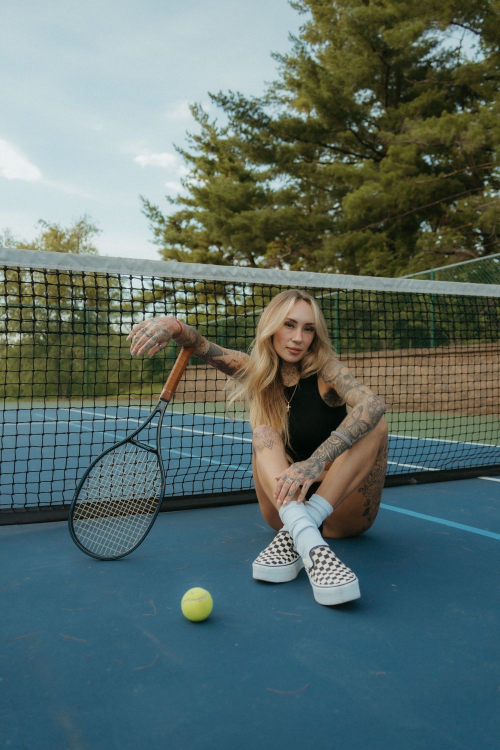 Girl posing on a tennis court while holding a tennis ball, with a racket nearby and the court's net and markings visible