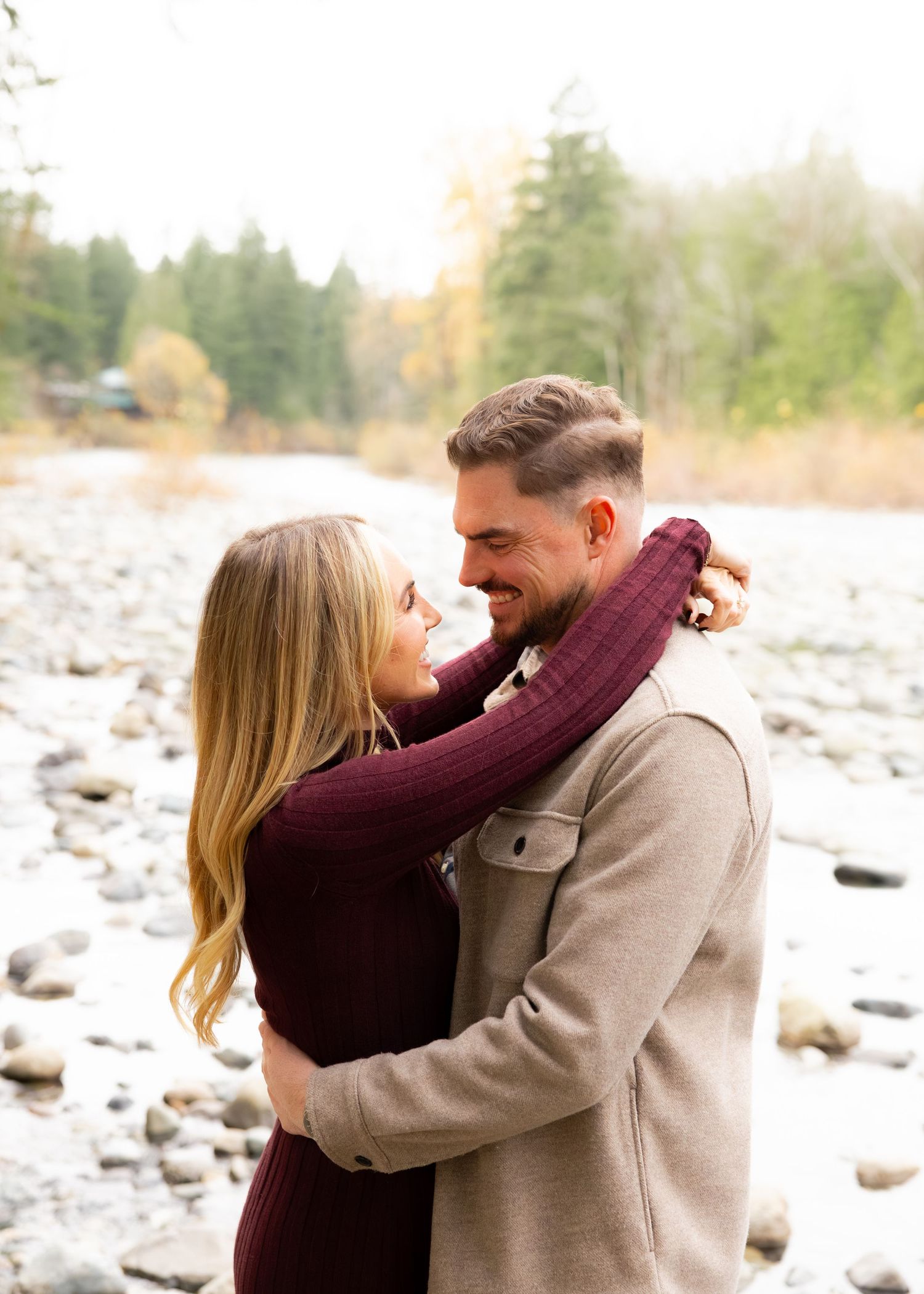 Romantic couple embracing by rocky riverbed with mountain and forest backdrop.