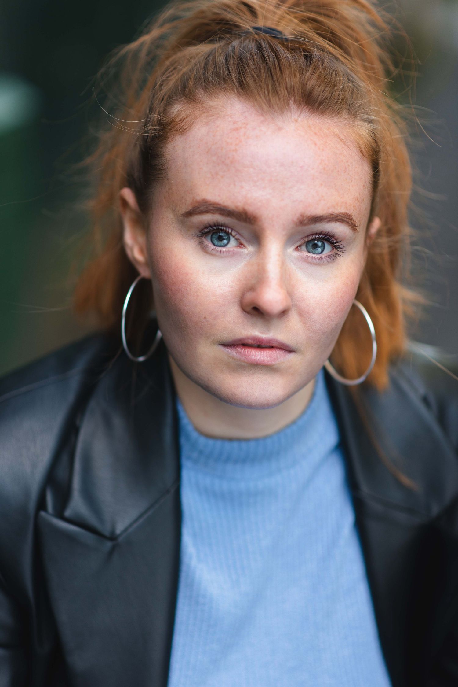 Arts Ed graduate wearing a blue sweater and leather jacket with large hoop earrings against a blurred background during headshot photoshoot