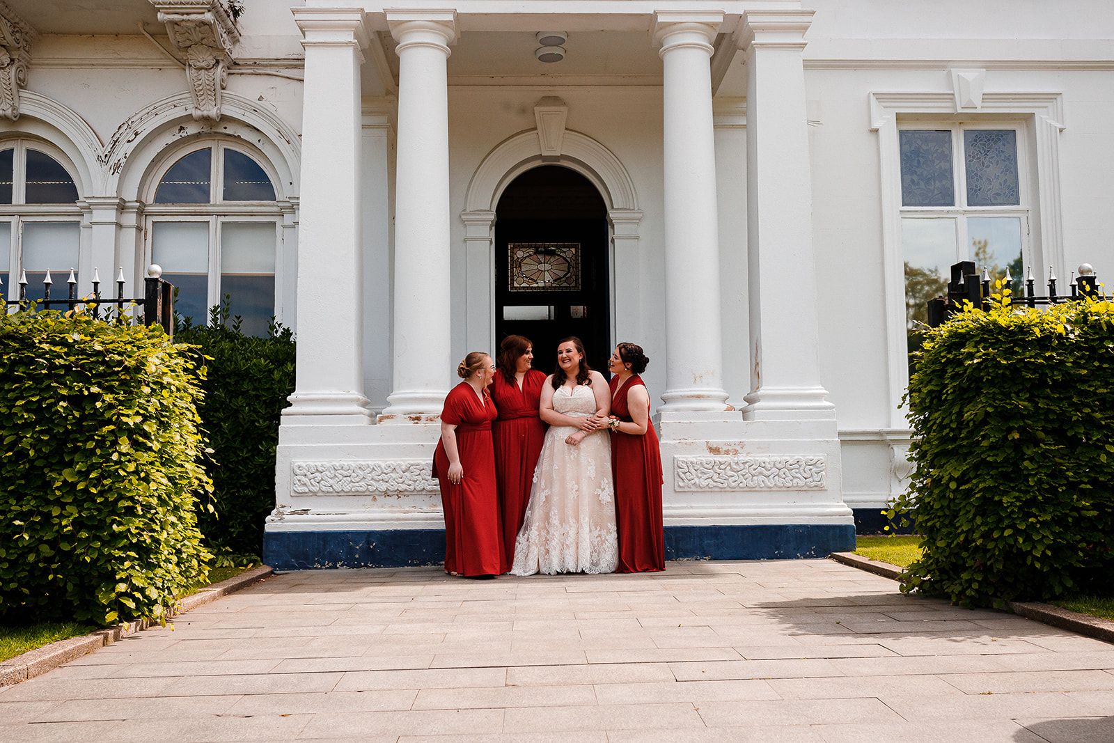 Bride in white gown poses with three bridesmaids in red dresses outside a grand white colonial building.