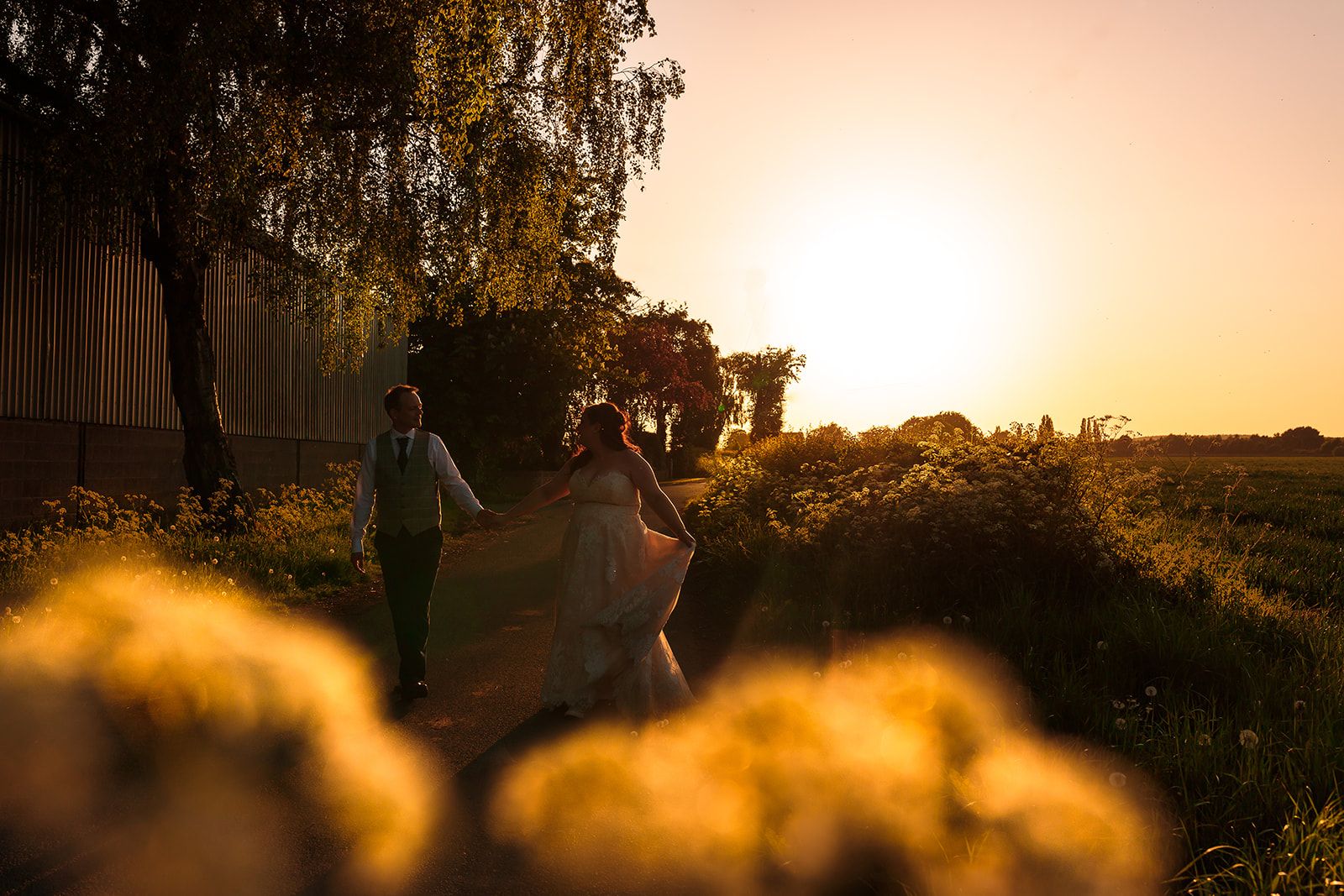 Couple walking hand in hand at golden sunset, surrounded by wildflowers and trees, in a romantic outdoor wedding setting.