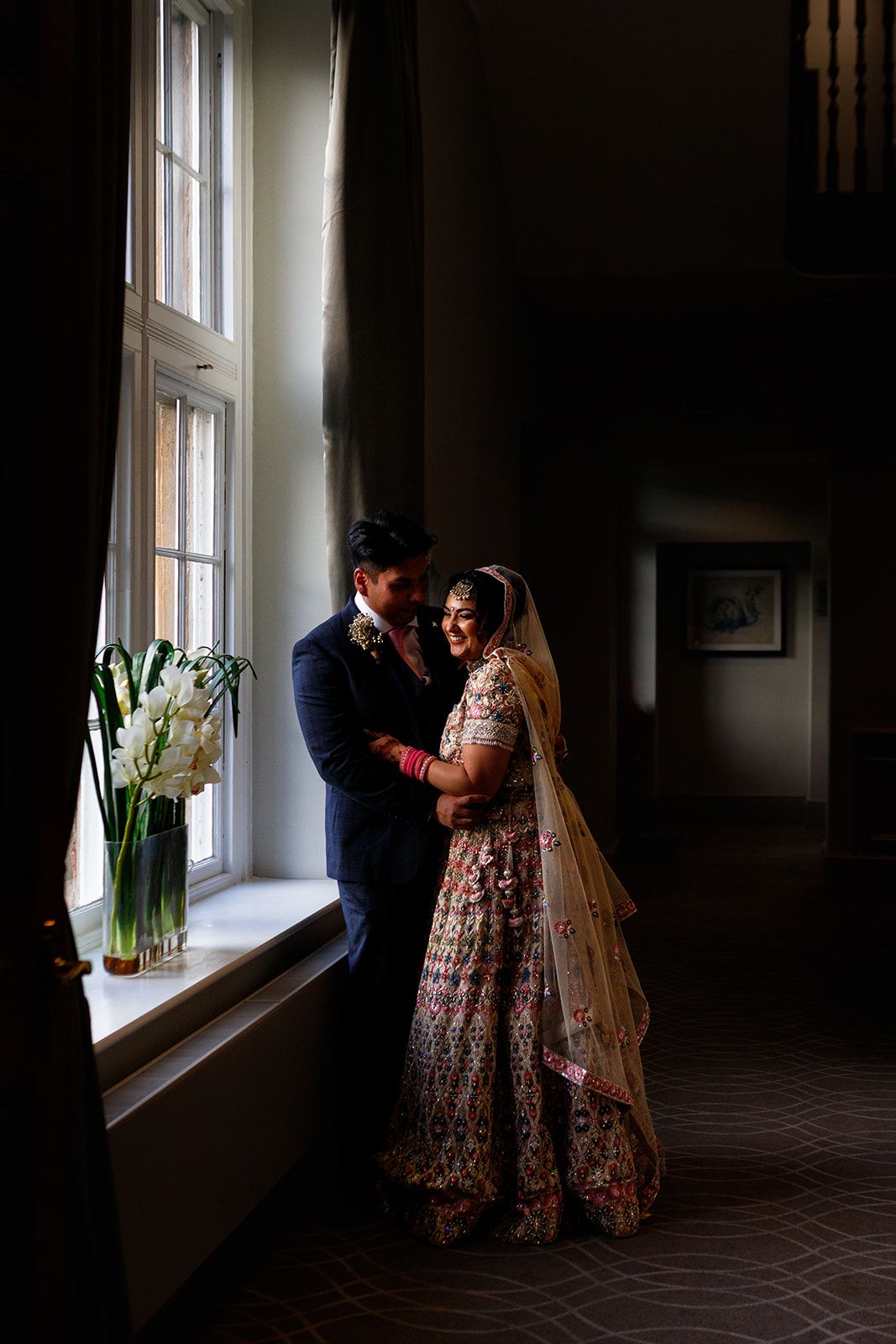 Indian bride in floral lehenga and groom in navy suit share romantic moment by sunlit window at wedding venue.
