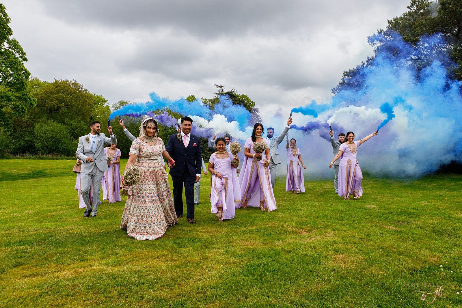 Wedding party walks on green lawn with blue and white smoke bombs, bride in gold gown, bridesmaids in pink dresses.
