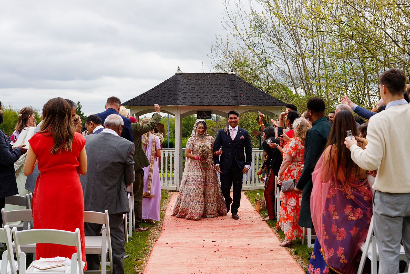 Bride and groom walk down outdoor wedding aisle lined with guests, gazebo backdrop, colorful attire.