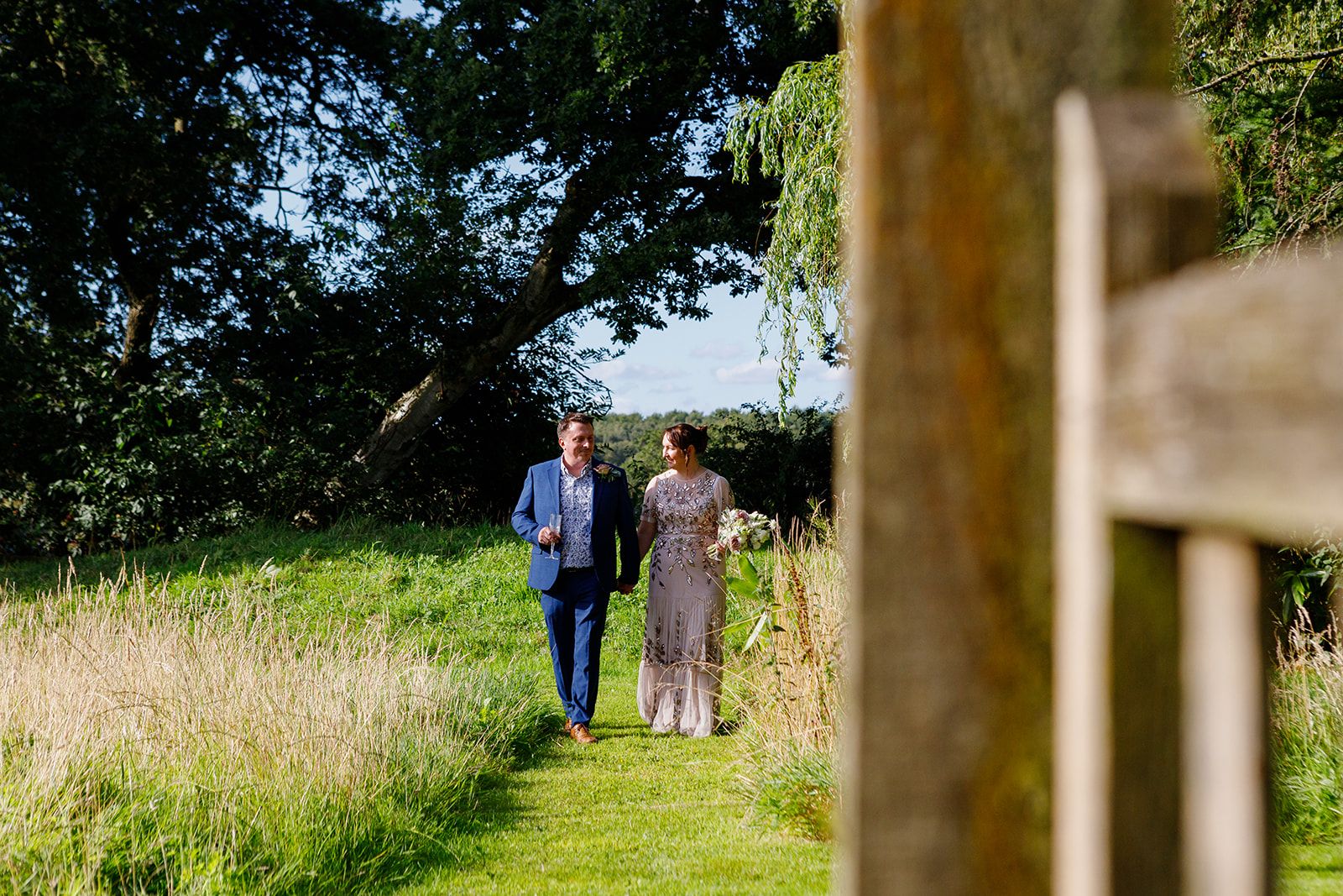 Couple walking together through a lush green countryside path surrounded by tall grass and trees on a sunny day.
