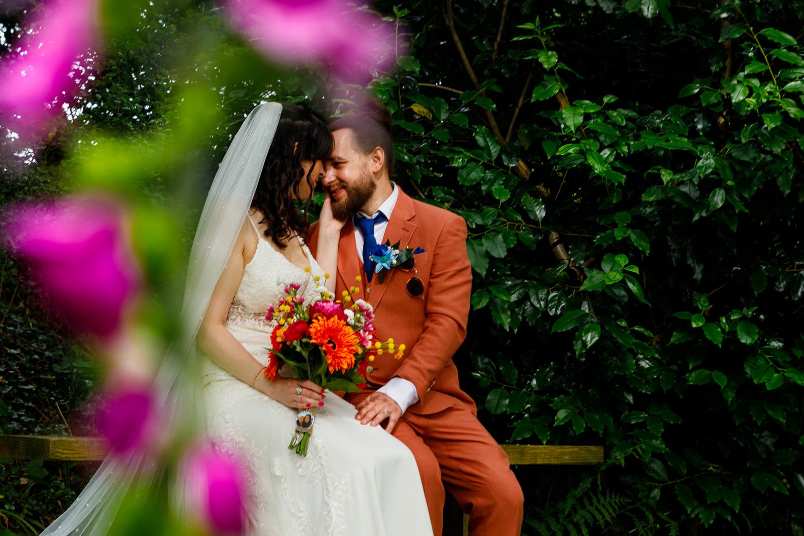 Bride and groom share a romantic moment surrounded by lush greenery, with vibrant pink flowers framing the couple.