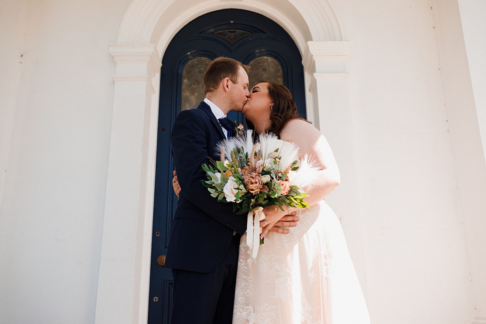 Bride and groom share a romantic kiss outside a church door, bride holding a colorful bouquet with protea flowers.