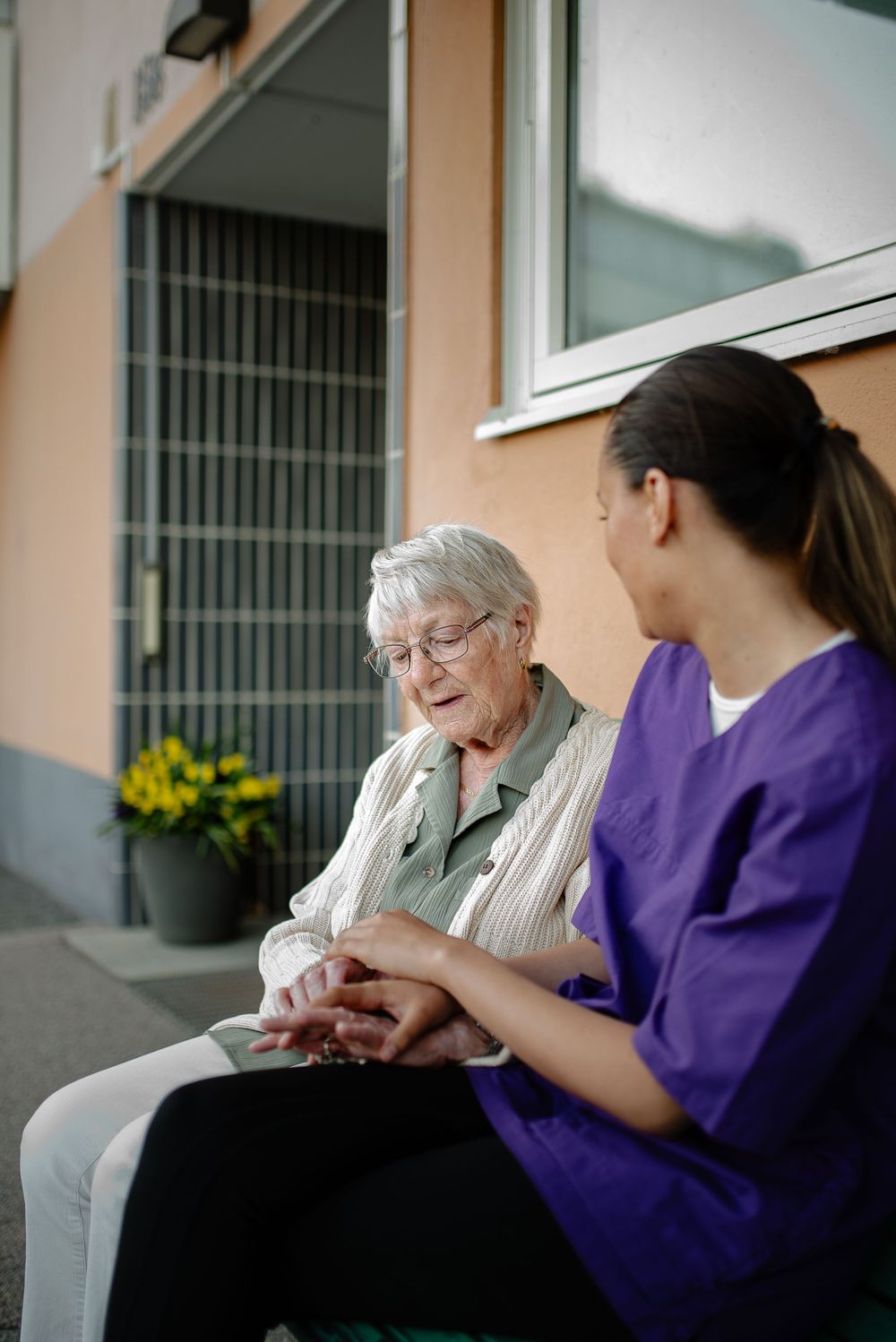A healthcare worker in purple scrubs provides companionship to an elderly resident outside.