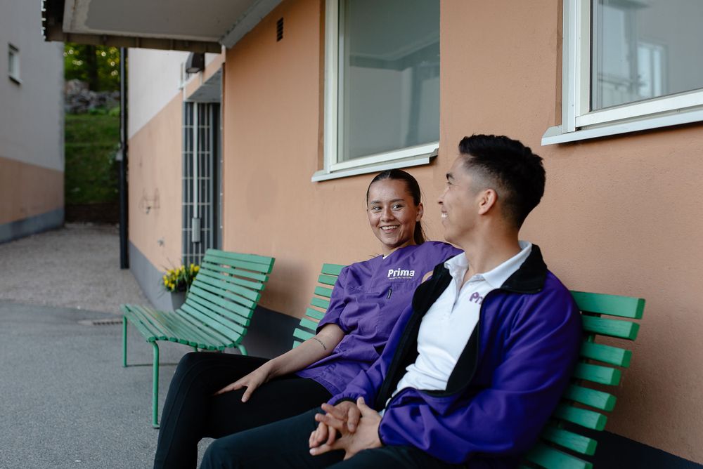 Two people share a relaxed moment while sitting on a green bench outside a building.