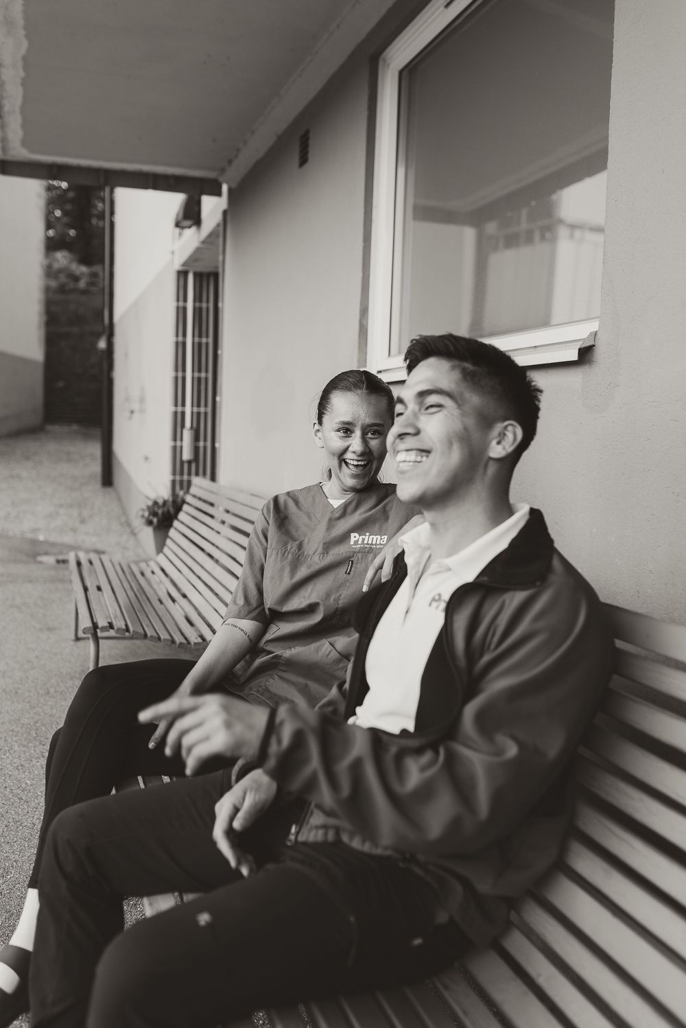 A candid black and white photo of two people sharing a laugh while sitting on an outdoor bench.