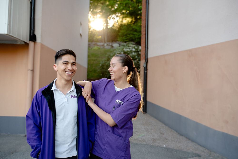 Two healthcare workers in purple uniforms share a lighthearted moment outside a medical facility at sunset.