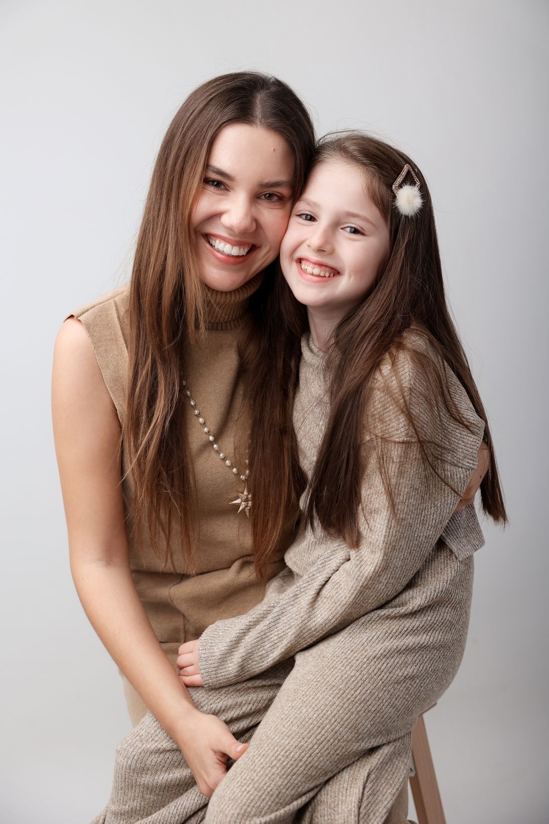 Portrait of two people wearing neutral-colored sweaters sharing a warm embrace against a light background.