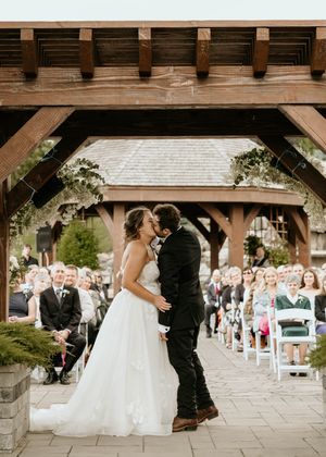 A couple shares a romantic moment during their outdoor wedding ceremony under a wooden gazebo with guests watching.