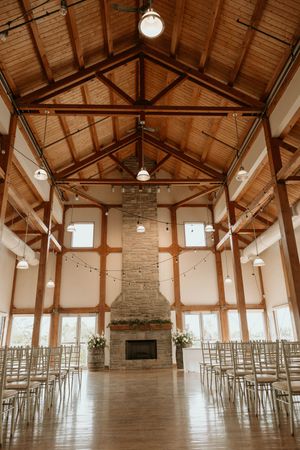 Rustic wedding venue interior with exposed wooden beams, stone fireplace and rows of white chairs in natural light.