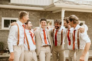 A cheerful wedding party of groomsmen in white shirts, khaki pants and red suspenders laugh together outdoors.