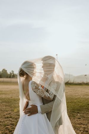 Romantic wedding couple embraces under flowing veil in sunlit field during golden hour sunset.