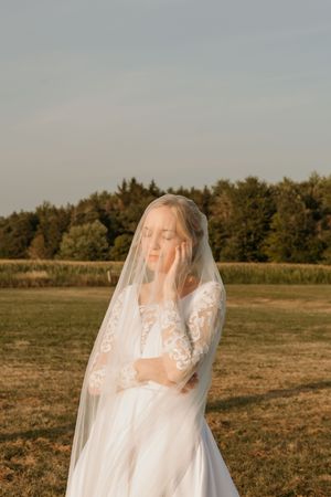 A figure in a flowing white wedding dress and veil stands gracefully in a sunlit field at golden hour.