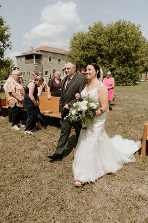 A beautiful outdoor wedding ceremony with guests gathered on wooden benches under trees on a sunny day.