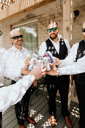 Groomsmen in white shirts and black vests toasting with beer cans at a rustic outdoor wedding venue.