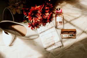Wedding details with red flowers, perfume bottle, invitation and shoes arranged in soft sunlight.