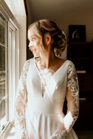 Bride in a classic white wedding dress with lace sleeves looks serenely toward window light in multiple poses.