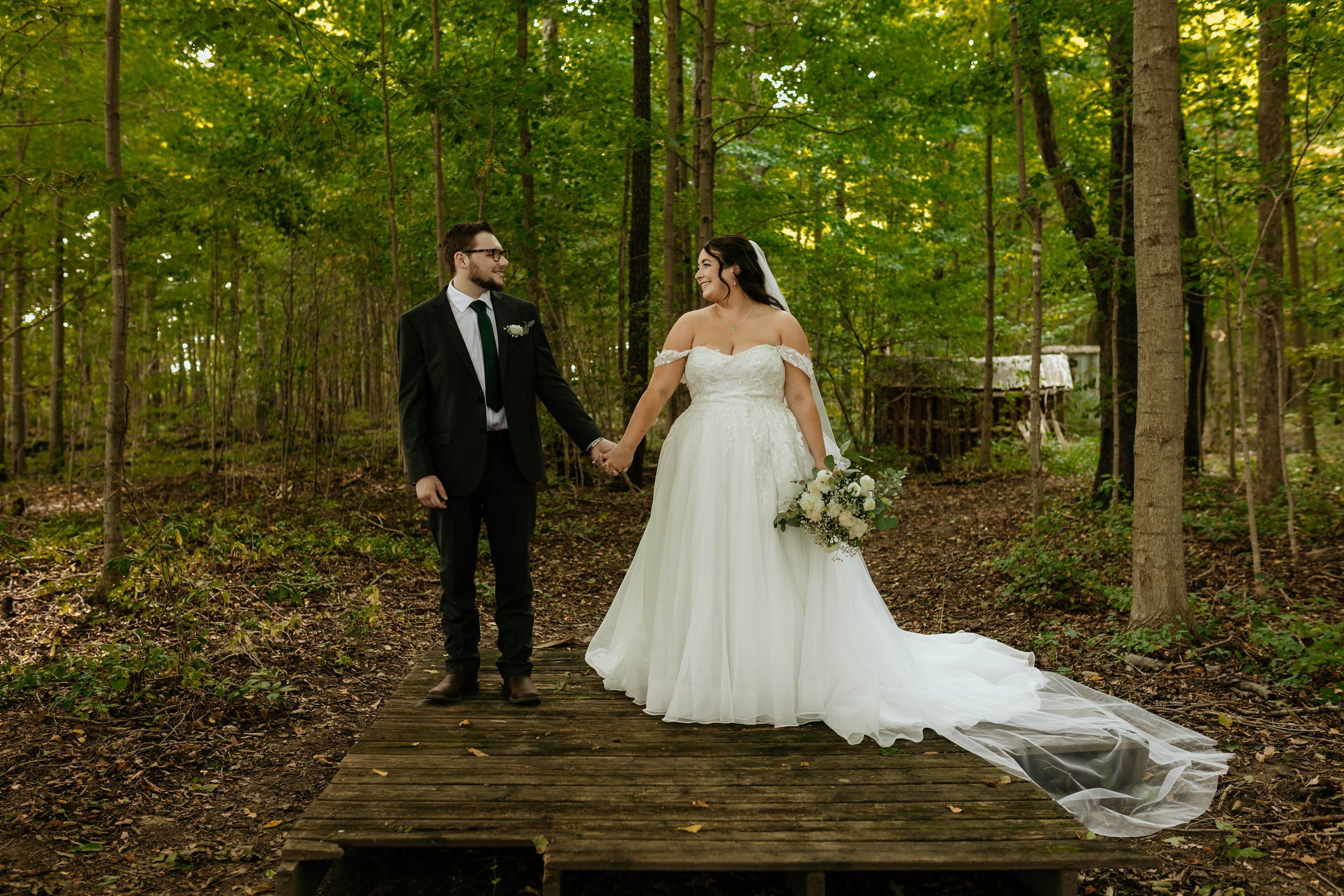 A wedding couple walks together on a wooden bridge path through a lush green forest setting.