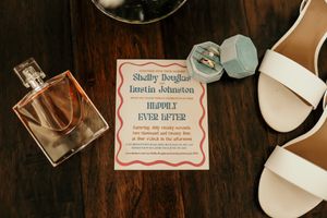 Wedding invitation with perfume bottle, bridal shoes, and greenery on dark wooden surface.