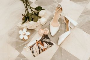 Wedding accessories including white heels, tulle veil, floral details and jewelry arranged on a checkered marble floor.