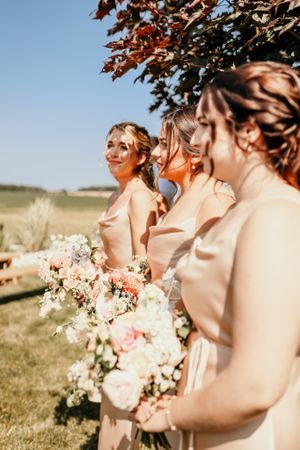 Bridesmaids in blush pink dresses hold elegant bouquets during an outdoor wedding ceremony.