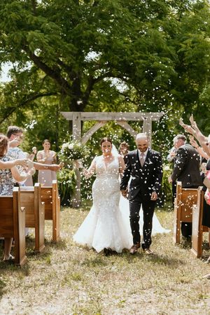 Wedding guests seated on wooden pews outdoors toss confetti as newlyweds celebrate under green trees.