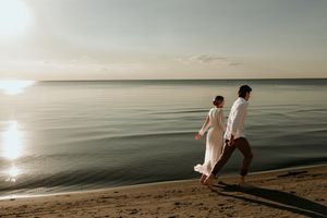 Two people in white clothing walking along a serene beachfront at sunset with calm waters in the background.